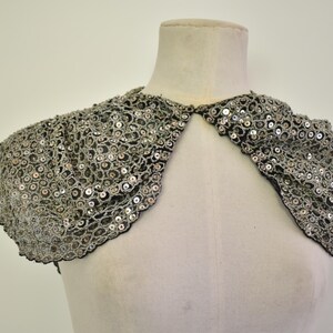 May include: A silver sequined bolero shrug featuring a delicate scalloped edge and intricate mesh detailing. The garment is designed with short, puffed sleeves and a front opening, showcasing a dense pattern of reflective sequins on a dark base.