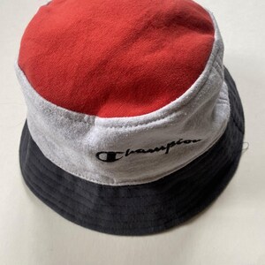 May include: A Champion bucket hat in a tri-colour design. The top is red, the middle section is grey, and the brim is black. The Champion logo is embroidered in black on the grey section.