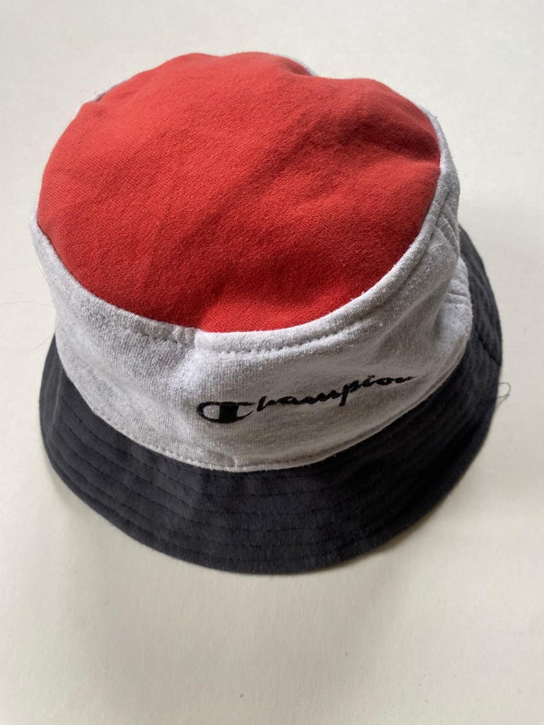 May include: A Champion bucket hat in a tri-colour design. The top is red, the middle section is grey, and the brim is black. The Champion logo is embroidered in black on the grey section.