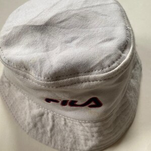 May include: A light grey bucket hat featuring the "FILA" logo in red and navy blue. The hat is made of a soft, textured material and has a classic, rounded shape. This hat is a casual accessory, suitable for various outfits.