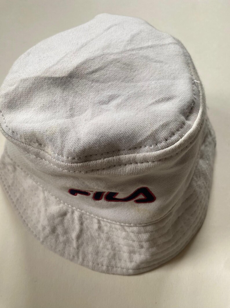 May include: A light grey bucket hat featuring the "FILA" logo in red and navy blue. The hat is made of a soft, textured material and has a classic, rounded shape. This hat is a casual accessory, suitable for various outfits.