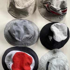 May include: A selection of six bucket hats in different colours and patterns. The hats include grey, black, and beige, some with brand logos and graphic details. The hats are made of soft, textured fabric.