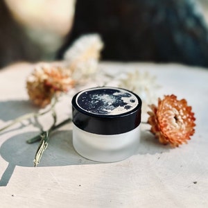 May include: A small, round jar with a black lid and a moon design on the lid. The jar is sitting on a wooden surface with dried flowers.