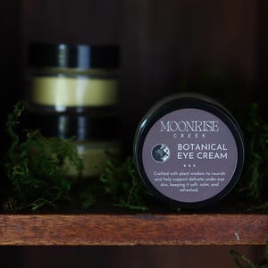 May include: A black jar of Moonrise Creek Botanical Eye Cream with a dark gray label, set in a wooden shelf with green moss. The label includes text about the product's benefits. Two additional jars are stacked behind it.