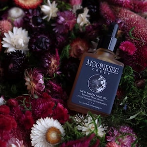 May include: A rectangular glass bottle of Moonrise Creek New Moon Face Oil with a black dropper cap, featuring a dark label with moon imagery and text, nestled among a dense arrangement of dried flowers in shades of red, pink, and white.