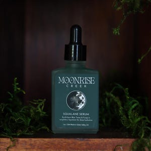 Könnte beinhalten: Eine teal-farbene Moonrise Creek Squalane Serum Flasche mit einer schwarzen Pipette. Die Flasche hat weißen Text und eine Mondgrafik. Die Flasche wird in einem Holzregal mit grünem Moos präsentiert.