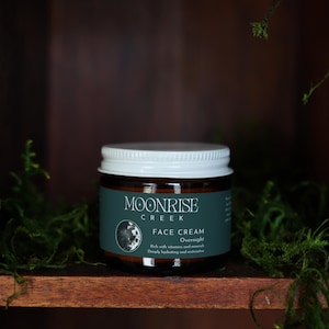 May include: A jar of Moonrise Creek Face Cream Overnight with a white lid and a dark green label. The jar is set in a wooden shelf with green moss.