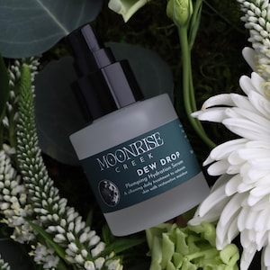 May include: A frosted glass bottle of Moonrise Creek Dew Drop Plumping Hydration Serum with a black pump top. The bottle is surrounded by white flowers, green foliage, and moss.