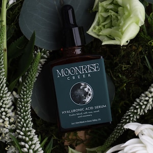 Op de afbeelding: Een bruine glazen fles Moonrise Creek Hyaluronzuur Serum met een zwarte druppelaar, geplaatst tegen een achtergrond van witte bloemen, groen gebladerte en mos. Het teal label toont een maan afbeelding en productdetails.