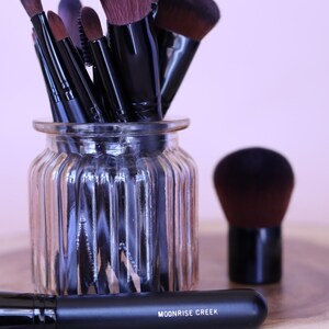 May include: A collection of makeup brushes with black handles and dark brown bristles, stored in a clear ribbed glass jar. One brush lies horizontally in the foreground with the text MOONRISE CREEK printed in white on its black handle.