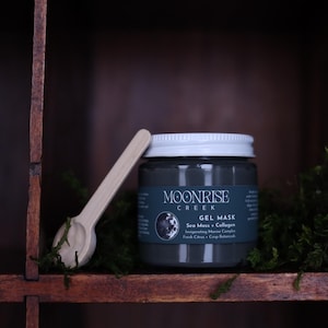 Pode incluir: Um frasco cinza-escuro de máscara em gel Moonrise Creek com tampa branca, apresentando um rótulo com o texto Moonrise Creek, Gel Mask, Sea Moss + Collagen, Invigorating Marine Complex, Fresh Citrus + Crisp Botanicals, e uma pequena espátula de madeira.