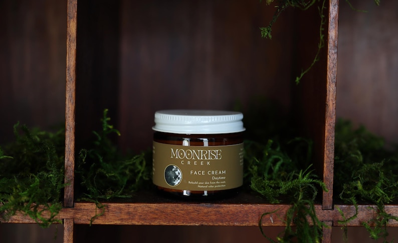 Puede incluir: Un tarro de crema facial Moonrise Creek, exhibido en una estanter&iacute;a de madera con musgo verde. El tarro tiene una tapa blanca y una etiqueta marr&oacute;n con el nombre de la marca y los detalles del producto. El fondo es una estructura de madera oscura.