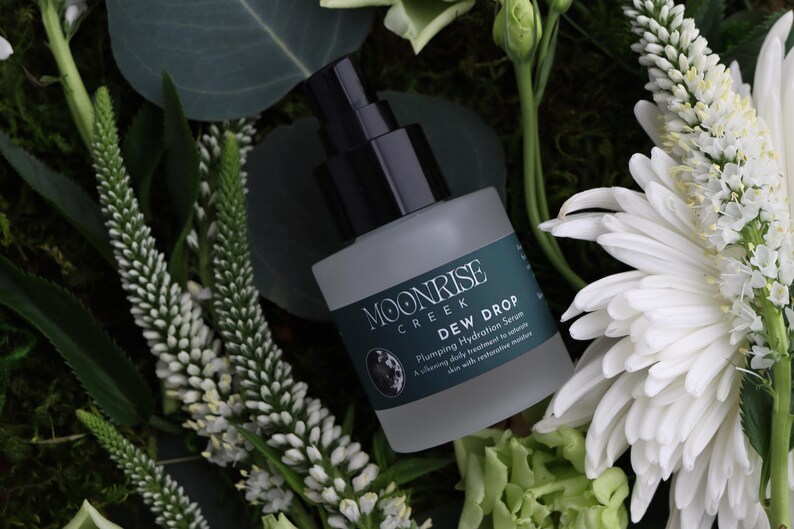 May include: A frosted glass bottle of Moonrise Creek Dew Drop Plumping Hydration Serum with a black pump top. The bottle is surrounded by white flowers, green foliage, and moss.
