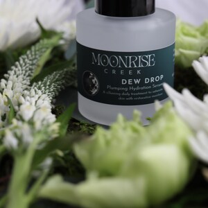 May include: A frosted glass bottle of Moonrise Creek Dew Drop Plumping Hydration Serum sits amidst white flowers and green foliage. The bottle has a black cap and a dark green label with white text. The serum is a daily treatment to saturate skin with restorative moisture.