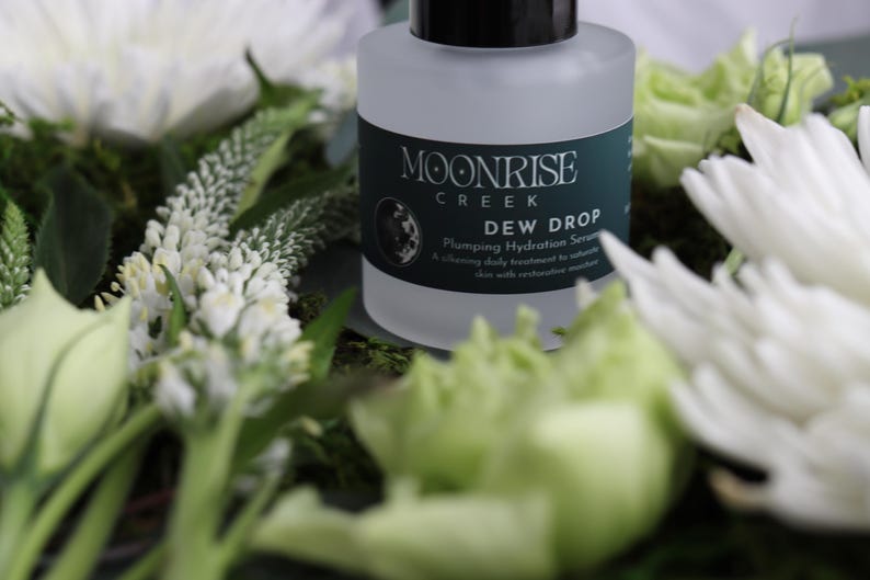 May include: A frosted glass bottle of Moonrise Creek Dew Drop Plumping Hydration Serum sits amidst white flowers and green foliage. The bottle has a black cap and a dark green label with white text. The serum is a daily treatment to saturate skin with restorative moisture.