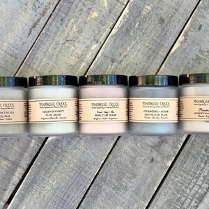 May include: Five glass jars of different colored clay masks. The jars are labeled with the names of the masks: Alchemy of the Sea, Green Divinity, Rose + Sage + Aloe Pink Clay Mask, Charcoal + Algae Detox Clay Mask, and Flower Child Moroccan Clay Mask.