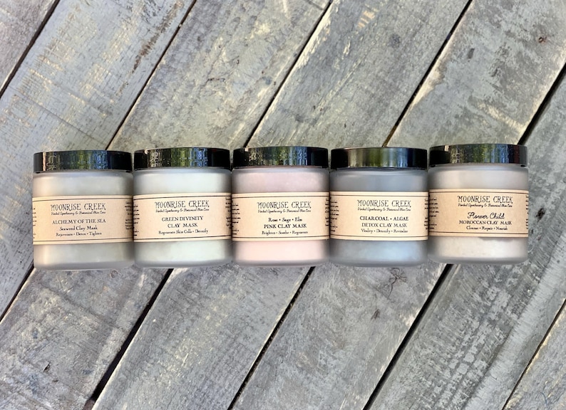 May include: Five glass jars of different colored clay masks. The jars are labeled with the names of the masks: Alchemy of the Sea, Green Divinity, Rose + Sage + Aloe Pink Clay Mask, Charcoal + Algae Detox Clay Mask, and Flower Child Moroccan Clay Mask.