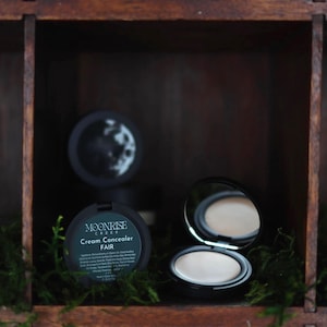 May include: A black compact of Moonrise Creek Cream Concealer in FAIR, with a mirror and a moon design on the lid. The compact is open, revealing the concealer. The product is displayed in a wooden box with green moss.