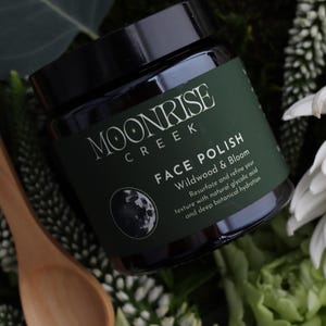 Op de afbeelding: Een pot Moonrise Creek Face Polish met een donkergroen etiket en een zwart deksel. Op het etiket staat "FACE POLISH Wildwood & Bloom". Een houten lepel en een witte bloem staan op de achtergrond.
