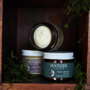 Puede incluir: Tres tarros de crema facial Moonrise Creek se exhiben en una estanter&iacute;a de madera. Un tarro est&aacute; abierto, revelando la crema. Los otros dos tarros est&aacute;n cerrados con tapas blancas. Las etiquetas dicen "Moonrise Creek Face Cream". Musgo verde se coloca alrededor de los tarros.