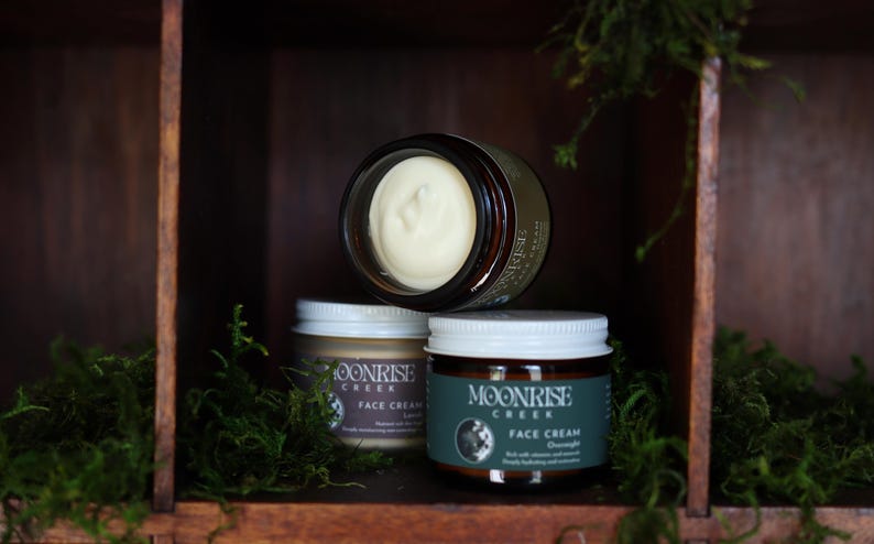 Puede incluir: Tres tarros de crema facial Moonrise Creek se exhiben en una estanter&iacute;a de madera. Un tarro est&aacute; abierto, revelando la crema. Los otros dos tarros est&aacute;n cerrados con tapas blancas. Las etiquetas dicen "Moonrise Creek Face Cream". Musgo verde se coloca alrededor de los tarros.