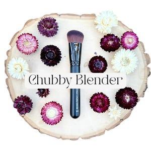 May include: A makeup brush with a black handle and dense, angled brown bristles. The handle features the text "chubby blender 160". The brush is surrounded by dried flowers in shades of deep purple, pink, and cream on a light wood slice.