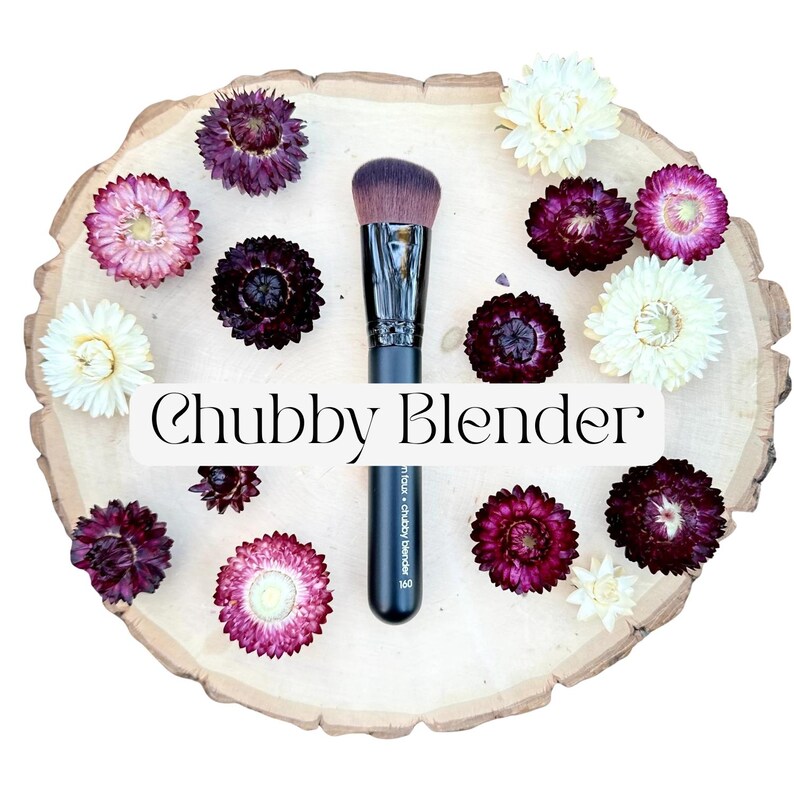May include: A makeup brush with a black handle and dense, angled brown bristles. The handle features the text "chubby blender 160". The brush is surrounded by dried flowers in shades of deep purple, pink, and cream on a light wood slice.