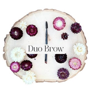 May include: A black dual-ended eyebrow brush with a spoolie on one end and an angled brush on the other, positioned vertically on a light-colored wooden slice surrounded by dried flowers in shades of white, pink, and deep purple, with the text Duo Brow.