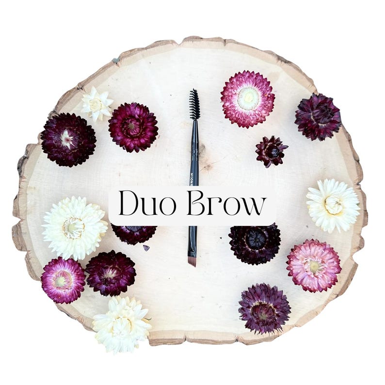 May include: A black dual-ended eyebrow brush with a spoolie on one end and an angled brush on the other, positioned vertically on a light-colored wooden slice surrounded by dried flowers in shades of white, pink, and deep purple, with the text Duo Brow.