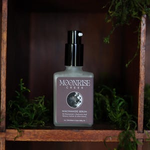 May include: A frosted glass bottle of Moonrise Creek Niacinamide Serum with a black pump dispenser. The label features the brand name, a moon graphic, and product information. The bottle is displayed on a wooden shelf with green moss.