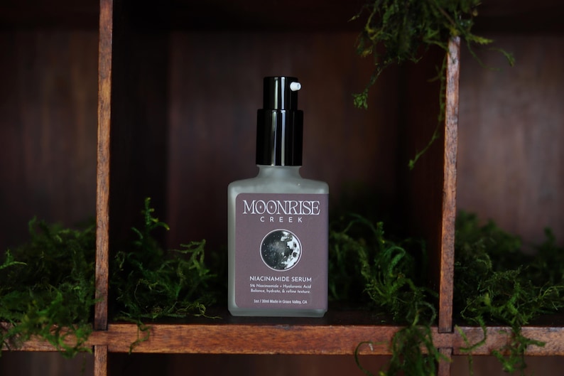 May include: A frosted glass bottle of Moonrise Creek Niacinamide Serum with a black pump dispenser. The label features the brand name, a moon graphic, and product information. The bottle is displayed on a wooden shelf with green moss.