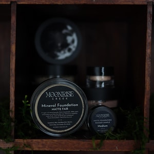 May include: A display of Moonrise Creek mineral foundation products. The image shows a large jar of Matte Fair foundation, a smaller sample, and other jars. The jars are black with white text and sit in a wooden shelf with greenery.