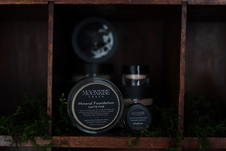 May include: A display of Moonrise Creek mineral foundation products. The image shows a large jar of Matte Fair foundation, a smaller sample, and other jars. The jars are black with white text and sit in a wooden shelf with greenery.