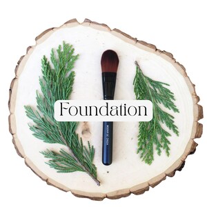 May include: A black foundation makeup brush with brown bristles, positioned vertically between two green cedar sprigs. The handle features the text "MOONRISE CREEK" in white, and a white rectangular label displays the word "Foundation".