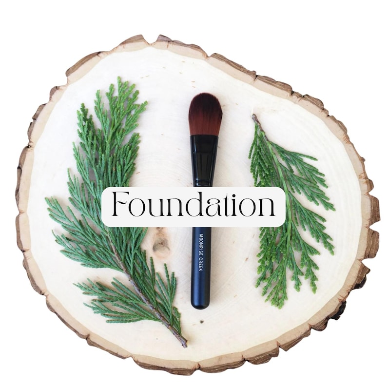 May include: A black foundation makeup brush with brown bristles, positioned vertically between two green cedar sprigs. The handle features the text "MOONRISE CREEK" in white, and a white rectangular label displays the word "Foundation".