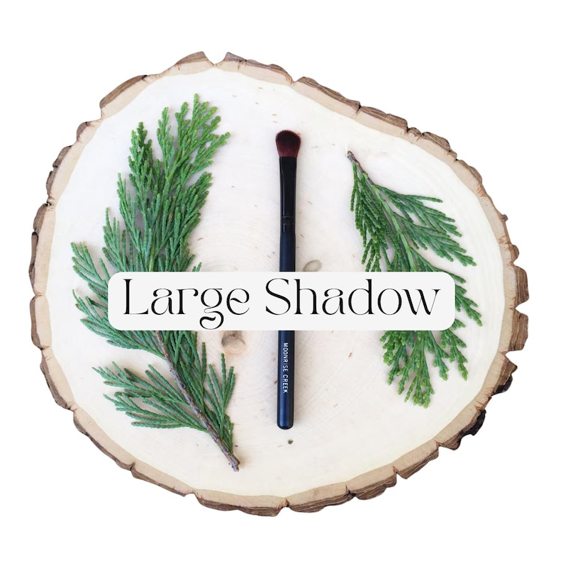 May include: A makeup brush with a black handle and dark bristles, featuring the text "MOONRISE CREEK" on the handle. The brush is positioned vertically between two green evergreen sprigs on a light-colored wood slice with the text "Large Shadow".