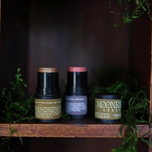 May include: Three cosmetic products are displayed on a wooden shelf with green moss. The products include two black tubes with gold and pink tops, and a small round container with the text "Moonrise Creek Sunrise Tints."