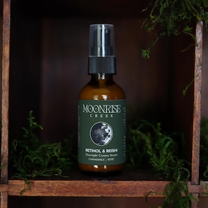 K&ouml;nnte beinhalten: Eine braune Glasflasche mit schwarzem Spr&uuml;hkopf, beschriftet mit "Moonrise Creek" und "Retinol & Reishi Overnight Creamy Serum". Die Flasche steht in einem Holzregal mit gr&uuml;nem Moos.