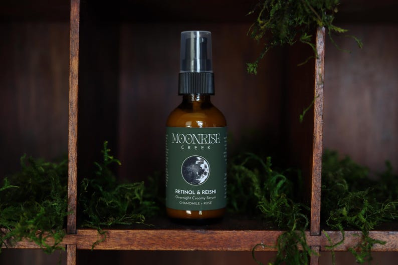 K&ouml;nnte beinhalten: Eine braune Glasflasche mit schwarzem Spr&uuml;hkopf, beschriftet mit "Moonrise Creek" und "Retinol & Reishi Overnight Creamy Serum". Die Flasche steht in einem Holzregal mit gr&uuml;nem Moos.