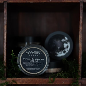May include: A jar of Moonrise Creek Mineral Foundation Matte Fair is displayed in a dark wooden cubby. The jar has a black label with white text. A decorative moon-themed lid is also visible. Other jars are in the background.