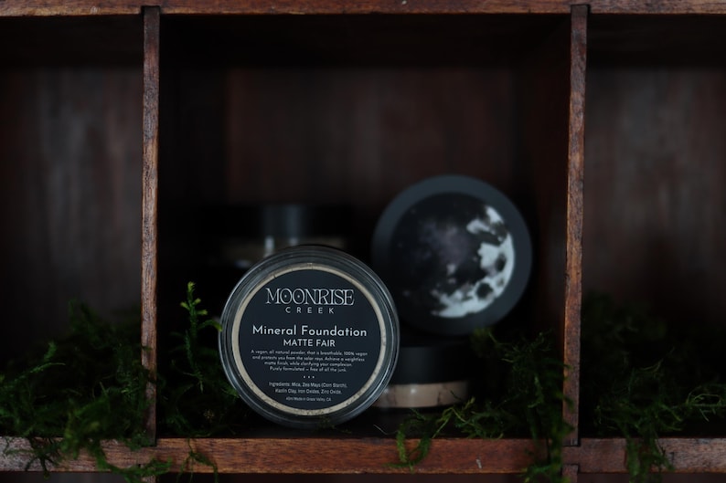 May include: A jar of Moonrise Creek Mineral Foundation Matte Fair is displayed in a dark wooden cubby. The jar has a black label with white text. A decorative moon-themed lid is also visible. Other jars are in the background.