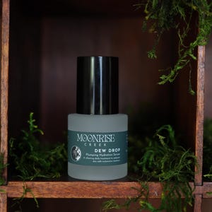 May include: A bottle of Moonrise Creek Dew Drop serum with a black cap and a gray label. The label includes the brand name, product name, and a description. The bottle is set against a wooden shelf with green moss.