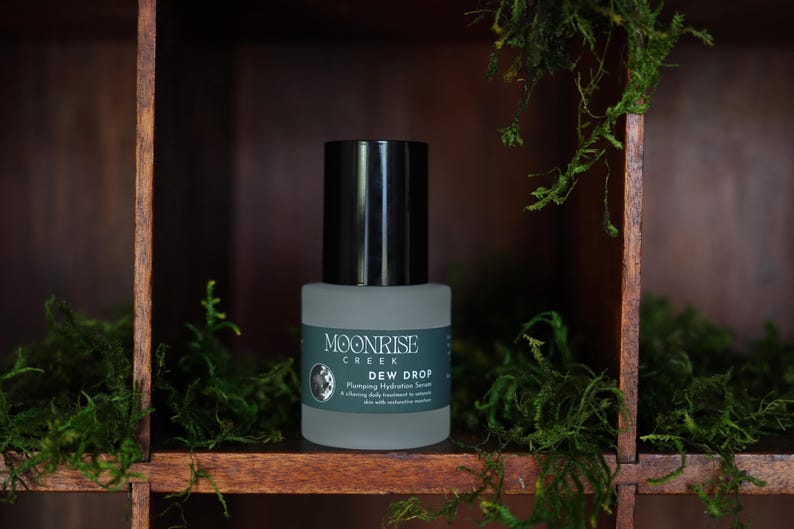 May include: A bottle of Moonrise Creek Dew Drop serum with a black cap and a gray label. The label includes the brand name, product name, and a description. The bottle is set against a wooden shelf with green moss.