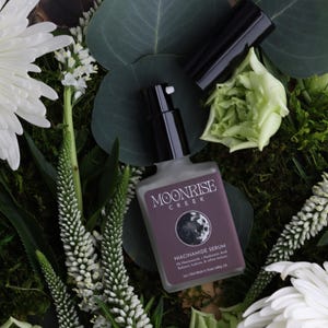 May include: A bottle of Moonrise Creek Niacinamide Serum with a black pump and cap, surrounded by white flowers, green leaves, and moss. The label is purple with white text.