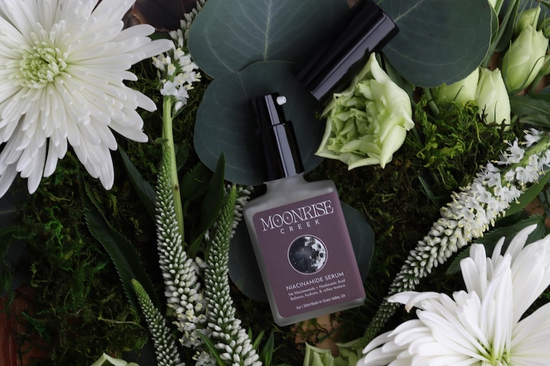 May include: A bottle of Moonrise Creek Niacinamide Serum with a black pump and cap, surrounded by white flowers, green leaves, and moss. The label is purple with white text.