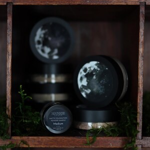 May include: A wooden shelf displays several black containers of Moonrise Matte Foundation Powder Sample. The containers have a moon design on the lids. The word "Medium" is visible on one container. Green moss is in the foreground.