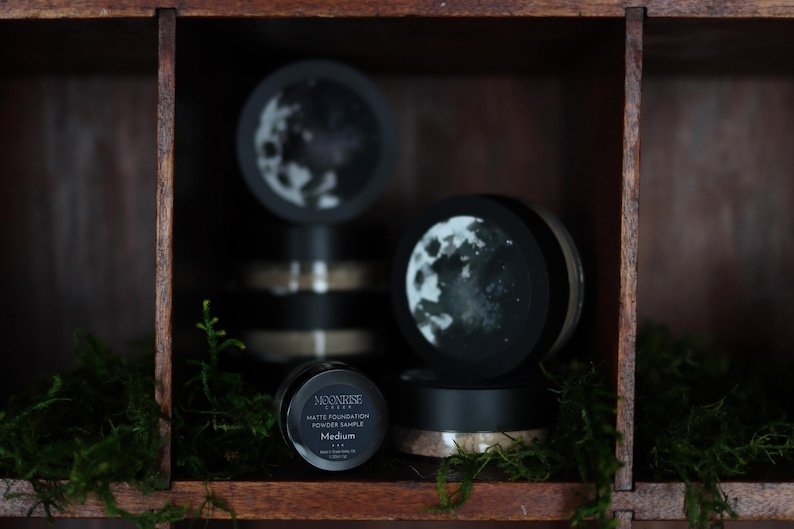 May include: A wooden shelf displays several black containers of Moonrise Matte Foundation Powder Sample. The containers have a moon design on the lids. The word "Medium" is visible on one container. Green moss is in the foreground.