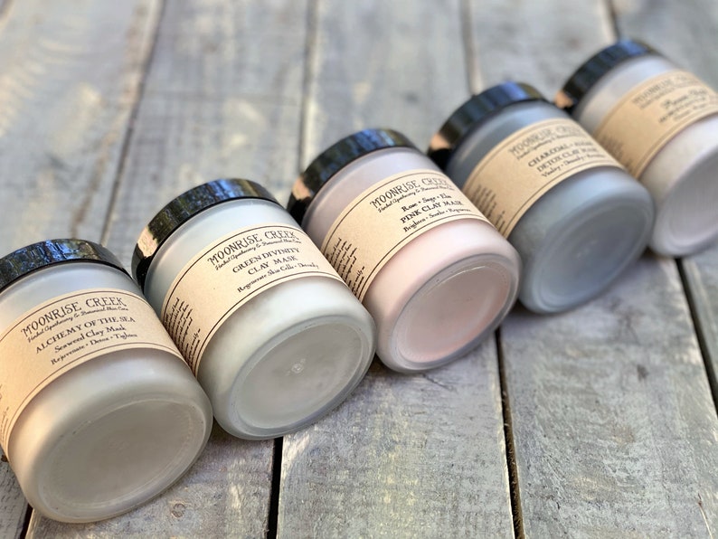 May include: Five glass jars with black lids containing different types of clay masks. The labels on the jars read "Moonrise Creek", "Alchemy of the Sea Seaweed Clay Mask", "Green Divinity Clay Mask", "Rose & Clay Pink Clay Mask", and "Charcoal Detox Clay Mask".