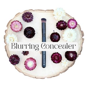 May include: A black makeup brush with a flat, dense head and a handle labeled Blurring Concealer 082, positioned vertically on a light-colored wooden slice surrounded by dried flowers in shades of deep purple, pink, and cream white.