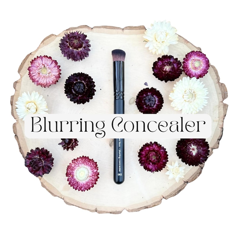 May include: A black makeup brush with a flat, dense head and a handle labeled Blurring Concealer 082, positioned vertically on a light-colored wooden slice surrounded by dried flowers in shades of deep purple, pink, and cream white.